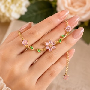 May include: A gold-tone chain bracelet featuring three pink floral motifs made of marquise-cut crystals. The design includes small green marquise-cut crystal leaves along the vine-like chain and a dangling pink flower charm at the end of the extender.