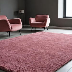 May include: A rectangular, solid dusty rose area rug with a thick, textured pile surface. The rug features a uniform, deep pink colour throughout its entire expanse, providing a soft and plush appearance that covers the floor space in the room.