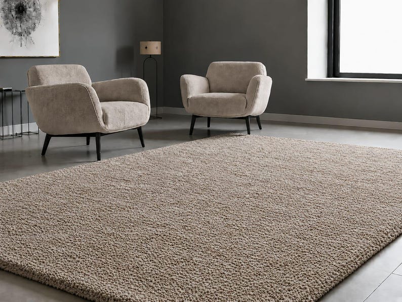 May include: A large rectangular area rug with a thick, textured, beige pile surface. The rug features a dense, looped construction and is positioned in a room with two matching beige upholstered armchairs with black wooden legs.