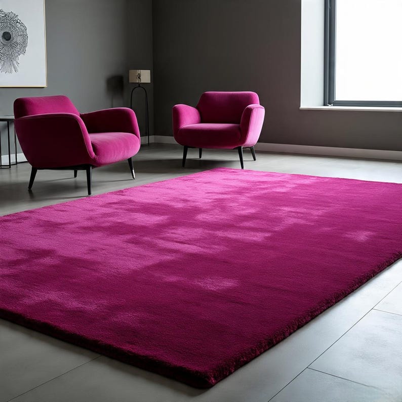 May include: A large, rectangular, deep magenta area rug with a plush, soft-textured surface. The rug is positioned on a smooth, light grey floor in front of two matching magenta upholstered armchairs with dark, slender legs.