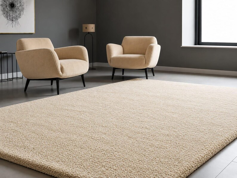 May include: A large, rectangular, cream-coloured area rug with a thick, textured, high-pile surface. The rug is positioned in the foreground, covering the floor in front of two matching cream-coloured armchairs with dark, tapered wooden legs.