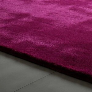 May include: A plush, deep magenta area rug with a dense, soft-textured pile. The rug features a uniform, solid colour throughout its surface, showcasing subtle variations in light and shadow across the fibres, creating a rich, velvety appearance.