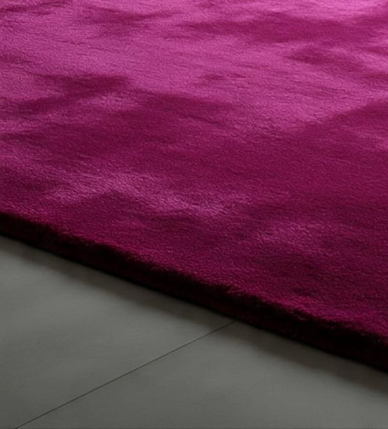 May include: A plush, deep magenta area rug with a dense, soft-textured pile. The rug features a uniform, solid colour throughout its surface, showcasing subtle variations in light and shadow across the fibres, creating a rich, velvety appearance.