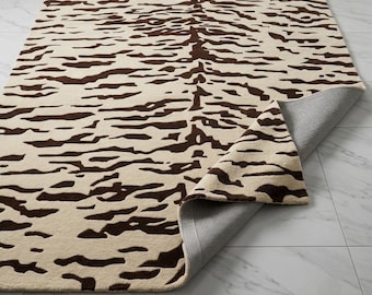 May include: A rectangular area rug featuring a cream-colored background with a dark brown abstract tiger stripe pattern. One corner of the rug is folded over to reveal a gray, textured backing material underneath the soft pile surface.
