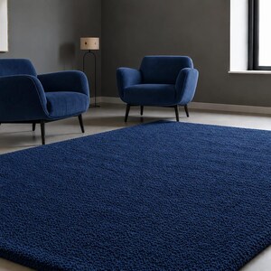 May include: A large, rectangular, deep blue area rug with a thick, textured, shaggy pile surface. The rug is positioned in the foreground of a room that contains two matching deep blue upholstered armchairs with dark wooden legs.