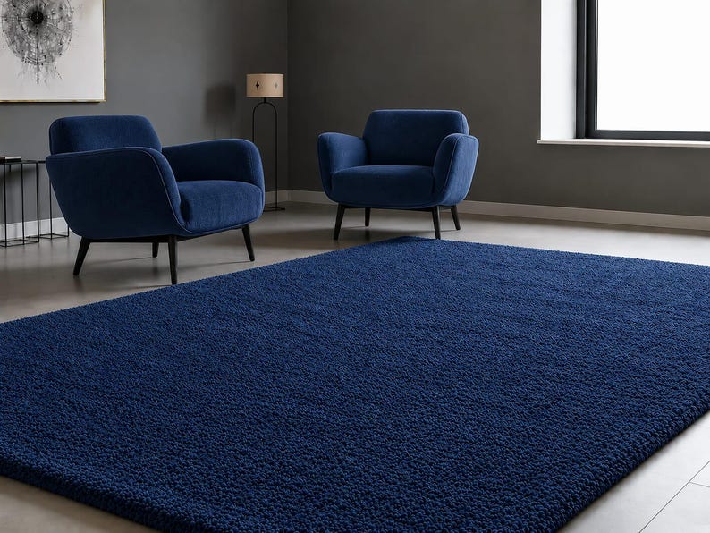 May include: A large, rectangular, deep blue area rug with a thick, textured, shaggy pile surface. The rug is positioned in the foreground of a room that contains two matching deep blue upholstered armchairs with dark wooden legs.