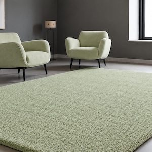 May include: A rectangular area rug with a thick, textured, light sage green pile. The rug features a dense, soft-touch surface and is positioned in a room with two matching light sage green armchairs with black legs and a dark grey wall.