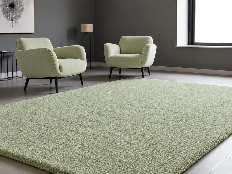 May include: A rectangular area rug with a thick, textured, light sage green pile. The rug features a dense, soft-touch surface and is positioned in a room with two matching light sage green armchairs with black legs and a dark grey wall.
