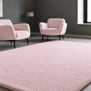 May include: A rectangular area rug with a soft, textured, light pink pile surface. The rug is positioned on a smooth grey floor in front of two matching light pink upholstered armchairs with black legs, set against a dark grey wall.