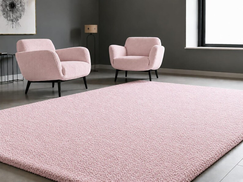 May include: A rectangular area rug with a soft, textured, light pink pile surface. The rug is positioned on a smooth grey floor in front of two matching light pink upholstered armchairs with black legs, set against a dark grey wall.