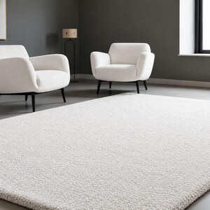 May include: A large, rectangular, off-white area rug with a dense, textured pile surface. The rug is positioned on a smooth grey floor in front of two matching off-white armchairs that feature dark, tapered wooden legs and curved backrests.