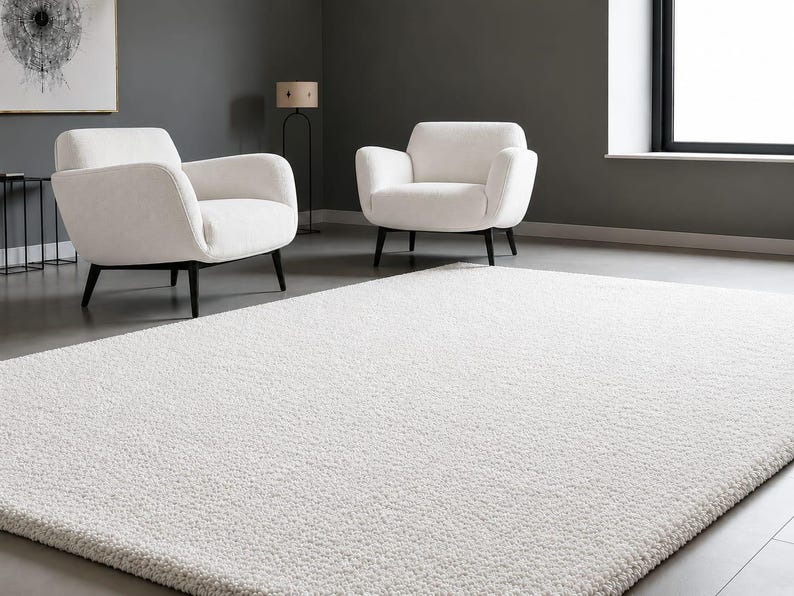 May include: A large, rectangular, off-white area rug with a dense, textured pile surface. The rug is positioned on a smooth grey floor in front of two matching off-white armchairs that feature dark, tapered wooden legs and curved backrests.