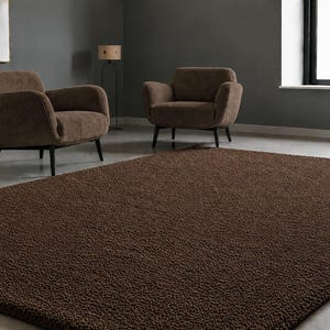 May include: A rectangular area rug featuring a deep brown colour and a dense, textured, shaggy pile surface. The rug is positioned on a smooth floor in front of two brown upholstered armchairs with dark wooden legs in a room with grey walls.