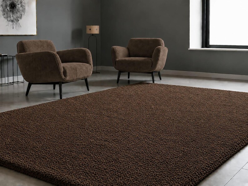 May include: A rectangular area rug featuring a deep brown colour and a dense, textured, shaggy pile surface. The rug is positioned on a smooth floor in front of two brown upholstered armchairs with dark wooden legs in a room with grey walls.