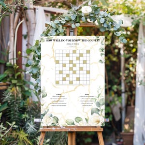 Può includere: Un cartello rettangolare per giochi di nozze intitolato How Well Do You Know The Couple? con un cruciverba, i nomi Anna & Marco e una data. Lo sfondo bianco presenta un motivo in marmo dorato, decorato con rose bianche ed eucalipto verde.