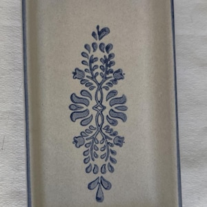 May include: A rectangular ceramic serving tray with a speckled off-white finish and a blue rim. The center of the tray features a symmetrical blue floral and leaf pattern oriented vertically, creating a clean and functional design for serving.