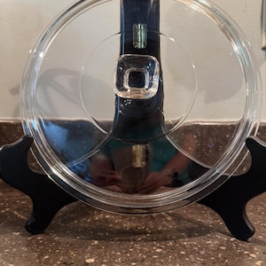 Könnte beinhalten: Ein runder, transparenter Glasdeckel für Kochgeschirr mit einem mittig platzierten, quadratischen Glasknauf als Griff. Der Deckel verfügt über einen glatten, leicht erhöhten Rand und eine klare, reflektierende Oberfläche für Kochtöpfe.