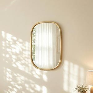 Op de afbeelding: Een wandspiegel met een glad, afgerond rechthoekig frame in een metallic gouden afwerking. Het reflecterende glazen oppervlak toont een raam met witte gordijnen, terwijl het frame zachte, gebogen hoeken en een eenvoudig ontwerp heeft.