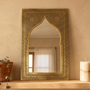 Peut inclure: Un miroir rectangulaire en laiton avec un sommet en arc et des motifs gravés sombres et complexes sur le cadre. La surface du miroir reflète une pièce avec une fenêtre et des rideaux, tandis que le bord en métal présente des motifs géométriques.