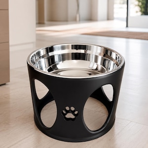 Pode incluir: Uma tigela elevada para animais de estimação com um recipiente de aço inoxidável removível inserido num suporte preto mate. O suporte tem uma base circular com grandes aberturas arqueadas e um design de pata recortado na parte frontal.