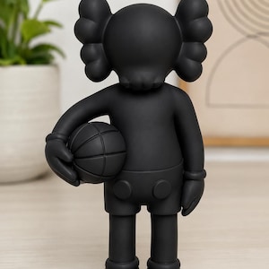 Può includere: Una statuetta da collezione in vinile nero opaco che raffigura un personaggio con occhi a forma di X e orecchie arrotondate, che tiene un pallone da basket nella mano destra. La figura ha una finitura nera liscia e indossa pantaloncini con due bottoni.