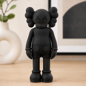 Può includere: Una statuetta da collezione nero opaco che raffigura un personaggio ispirato ai cartoni animati con orecchie arrotondate, occhi a forma di X e mani guantate. Il personaggio indossa pantaloncini con due bottoni circolari e grandi scarpe arrotondate.
