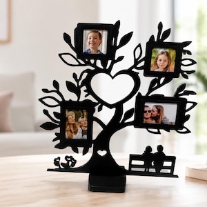Peut inclure: Un cadre photo en métal noir en forme d'arbre avec quatre emplacements rectangulaires, une découpe centrale en forme de cœur et la silhouette de deux personnages assis sur un banc à la base. Le cadre repose sur un socle rectangulaire.