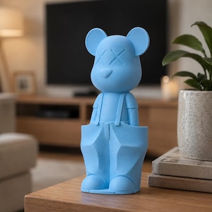 Peut inclure: Une figurine mate bleu clair représentant un personnage stylisé aux allures d'ours avec des oreilles rondes, un petit nez et des yeux en forme de X. Le personnage porte une salopette avec des bretelles et de grandes poches sur une base plate.