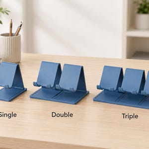 May include: Three blue plastic phone stands arranged in a row, labeled Single, Double, and Triple. Each stand features a slanted backrest and a small front lip to hold a device securely in place. The stands are matte blue and have a simple design.