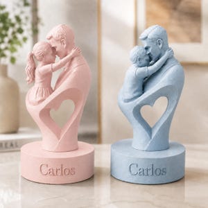 Pode incluir: Duas figuras esculpidas em bases circulares, uma rosa e outra azul. Cada uma retrata um adulto abraçando uma criança, formando um espaço vazio em forma de coração. O nome Carlos está gravado na frente de ambas as bases circulares.
