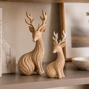 Könnte beinhalten: Zwei hellbeige Hirschfiguren mit glatten, geschwungenen, fließenden Linien, die in ihre Körper geschnitzt sind. Der größere Hirsch hat hohe, verzweigte Geweihe, während der kleinere kürzere, hell gespitzte Geweihe hat. Beide Figuren sitzen.