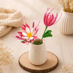 May include: Two crocheted flowers with pink, white, and yellow petals and long thin stamens, featuring green stems and leaves. The flowers are arranged in a small, ribbed, off-white ceramic vase that sits on a round wooden slice base.