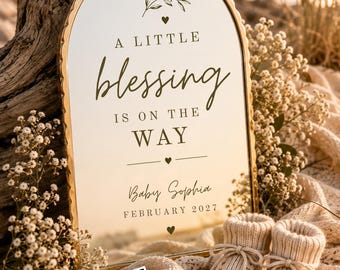 Op de afbeelding: Een boogvormige spiegel met een gouden geschulpte rand met de tekst A LITTLE blessing IS ON THE WAY, Baby Sophia en FEBRUARY 2027. Een paar beige gebreide babyslofjes en een echoscopie liggen voor de spiegel.