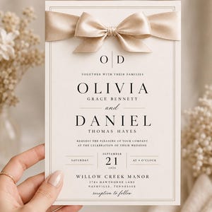 May include: A rectangular wedding invitation card featuring a cream background with a double-line border and a champagne-colored satin ribbon bow at the top. The text includes the names Olivia Grace Bennett and Daniel Thomas Hayes with event details.