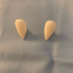 May include: Two off-white, pointed fangs, likely costume accessories, are displayed against a light blue fabric background. The fangs are symmetrical and appear to be made of a smooth material.