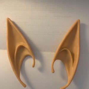 May include: A pair of prosthetic elf ears in a light skin tone. The ears are pointed and have a curved inner shape, designed to fit over the natural ear. Perfect for cosplay or costume parties.