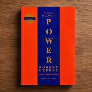 May include: A book titled The Concise 48 Laws of Power by Robert Greene, a Joost Elffers book, new edition. The cover features a vertical blue stripe centered on an orange background with gold text. A gold circular International Bestseller seal is visible.
