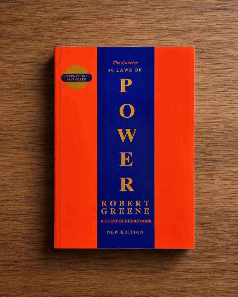 May include: A book titled The Concise 48 Laws of Power by Robert Greene, a Joost Elffers book, new edition. The cover features a vertical blue stripe centered on an orange background with gold text. A gold circular International Bestseller seal is visible.