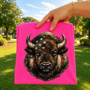 May include: A bright pink t-shirt featuring a detailed graphic of a bison head. The bison is rendered in shades of brown and black, with gold stars overlaid on its forehead and red and white stripes visible on the side of its face.