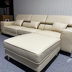 May include: A cream-colored leather sectional sofa with a matching ottoman. The sofa features adjustable headrests, three square throw pillows, and silver-toned metal accents on the armrests. The piece is supported by short, light-colored legs.