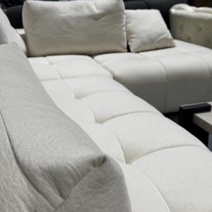 May include: A large off-white sectional sofa featuring a tufted seat design with square stitching patterns. The sofa includes multiple plush back cushions and a textured fabric upholstery, providing a wide seating area for multiple people.