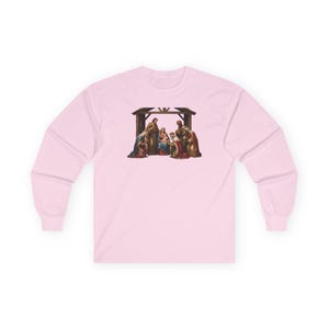 Puede incluir: Una camiseta de manga larga de color rosa claro con un estampado gráfico central de una escena de natividad. La ilustración muestra figuras en un establo, incluyendo un grupo central con un bebé, rodeado de personas con túnicas y coronas.