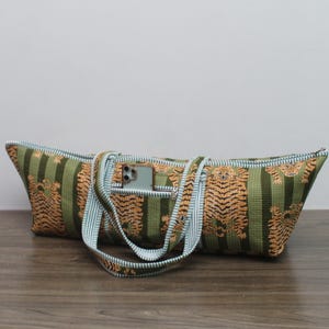 May include: A long, rectangular tote bag featuring a green and dark green striped pattern with an orange tiger print. The bag includes two light blue and white striped fabric handles and a small zippered exterior pocket containing a smartphone.