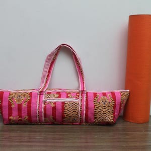 May include: A rectangular fabric yoga mat bag featuring a repeating tiger pattern on a background of vertical pink and red stripes. The bag includes two top handles and a central zippered pocket, positioned next to an orange rolled yoga mat.