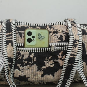 May include: A quilted fabric bag featuring a black and beige floral pattern with black and white striped straps. A light green smartphone with a butterfly sticker is tucked into the front zippered pocket of the bag, showing the camera lenses.
