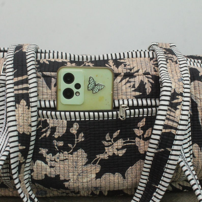 May include: A quilted fabric bag featuring a black and beige floral pattern with black and white striped straps. A light green smartphone with a butterfly sticker is tucked into the front zippered pocket of the bag, showing the camera lenses.
