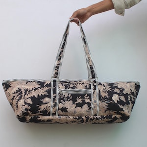 May include: A long, rectangular tote bag featuring a black and beige floral pattern. The bag has two long shoulder straps with a black and white striped design. A horizontal zippered pocket is located on the front exterior of the bag.