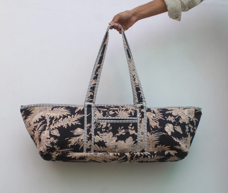 May include: A long, rectangular tote bag featuring a black and beige floral pattern. The bag has two long shoulder straps with a black and white striped design. A horizontal zippered pocket is located on the front exterior of the bag.