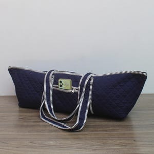 May include: A dark blue quilted tote bag featuring a diamond-stitched pattern and two long shoulder straps. The bag includes a small exterior zippered pocket on the front, which contains a light-colored smartphone with a visible camera lens.