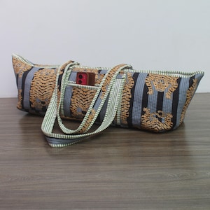 May include: A long, rectangular fabric tote bag featuring a repeating tiger pattern on a background of vertical black and light blue stripes. The bag has two matching fabric handles and a small exterior pocket containing a red rectangular object.